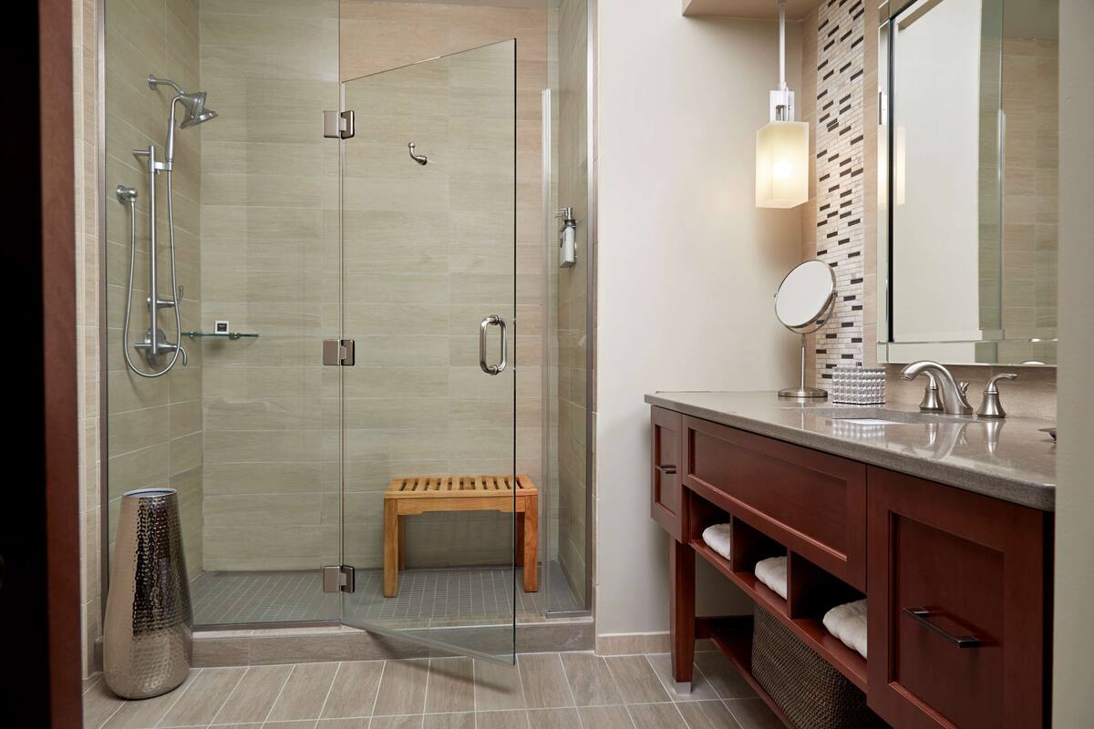 A modern bathroom with a glass-enclosed shower, a wooden bench, a vanity with a mirror, and a tiled floor.