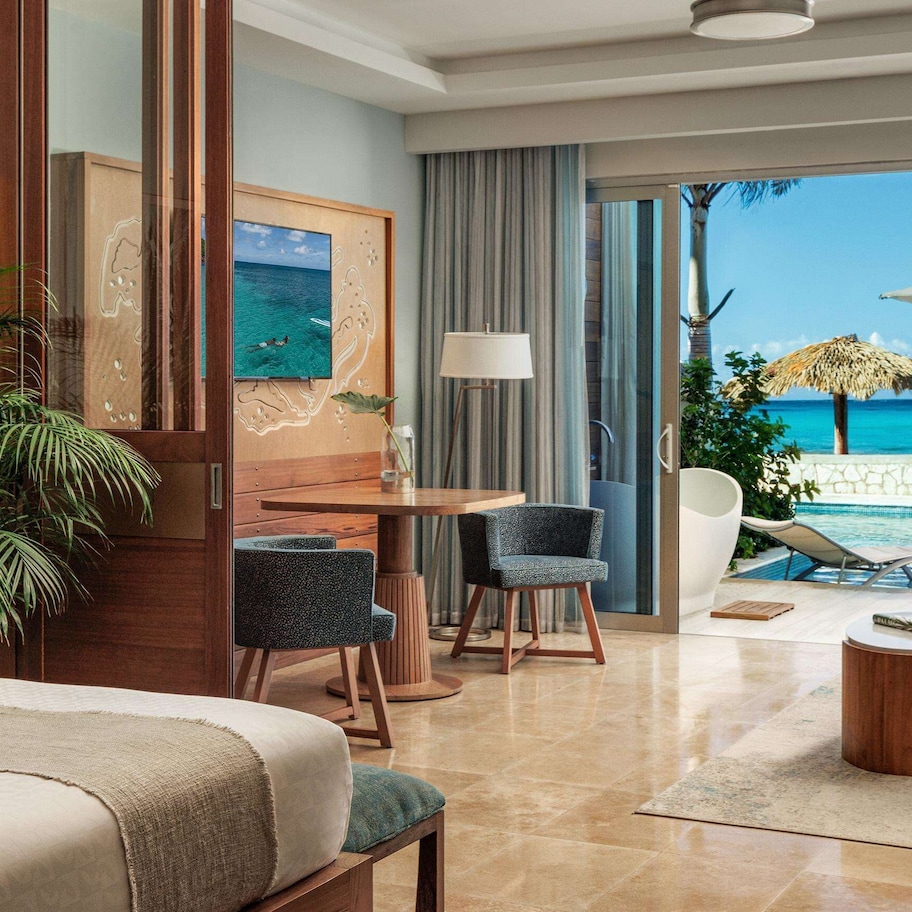 A hotel room with a bed, a desk, a chair, a small table, and a view of the beach and ocean.