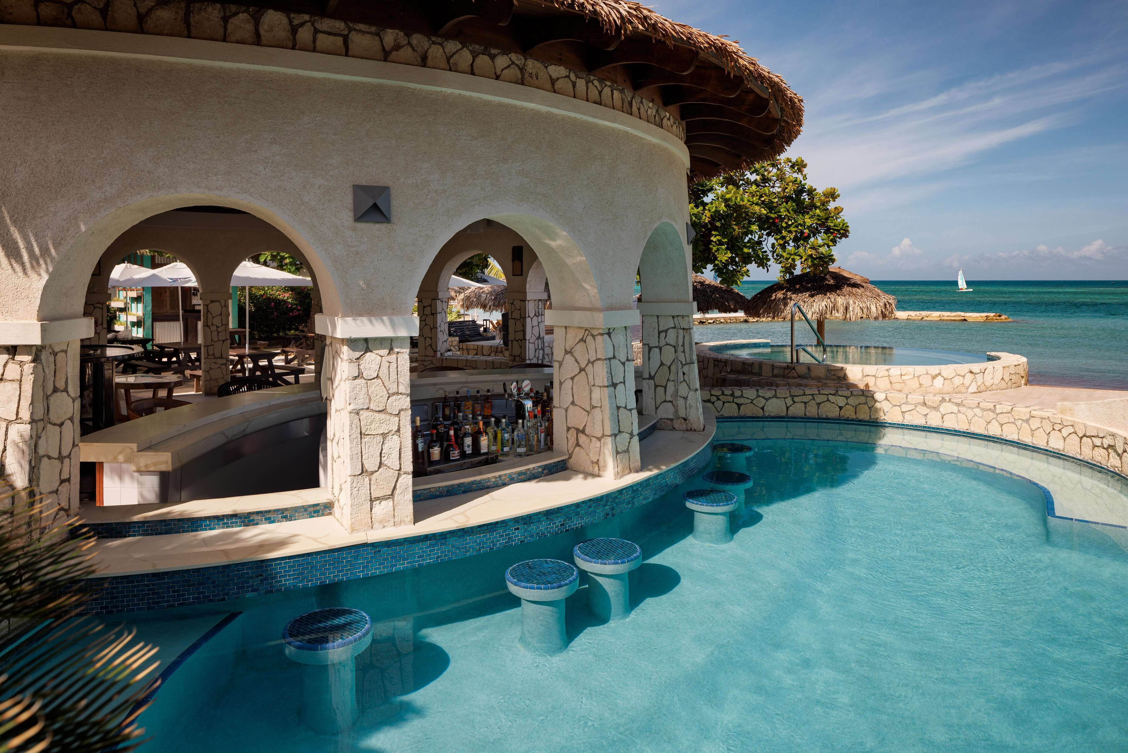 A poolside bar with stone arches, a bar counter, and a seating area with umbrellas.
