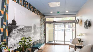 A reception area with a large wall mural, a bench, two chairs, a small table with a plant, and a door leading outside.