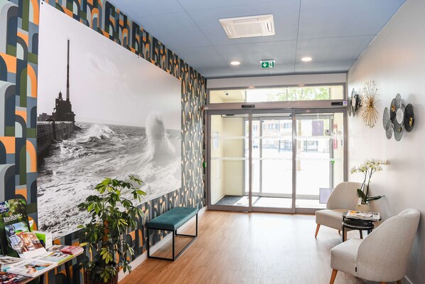 Un espace d’accueil agrémenté d’une grande fresque murale, d’un banc, de deux chaises, d’une petite table avec une plante et d’une porte donnant sur l’extérieur.