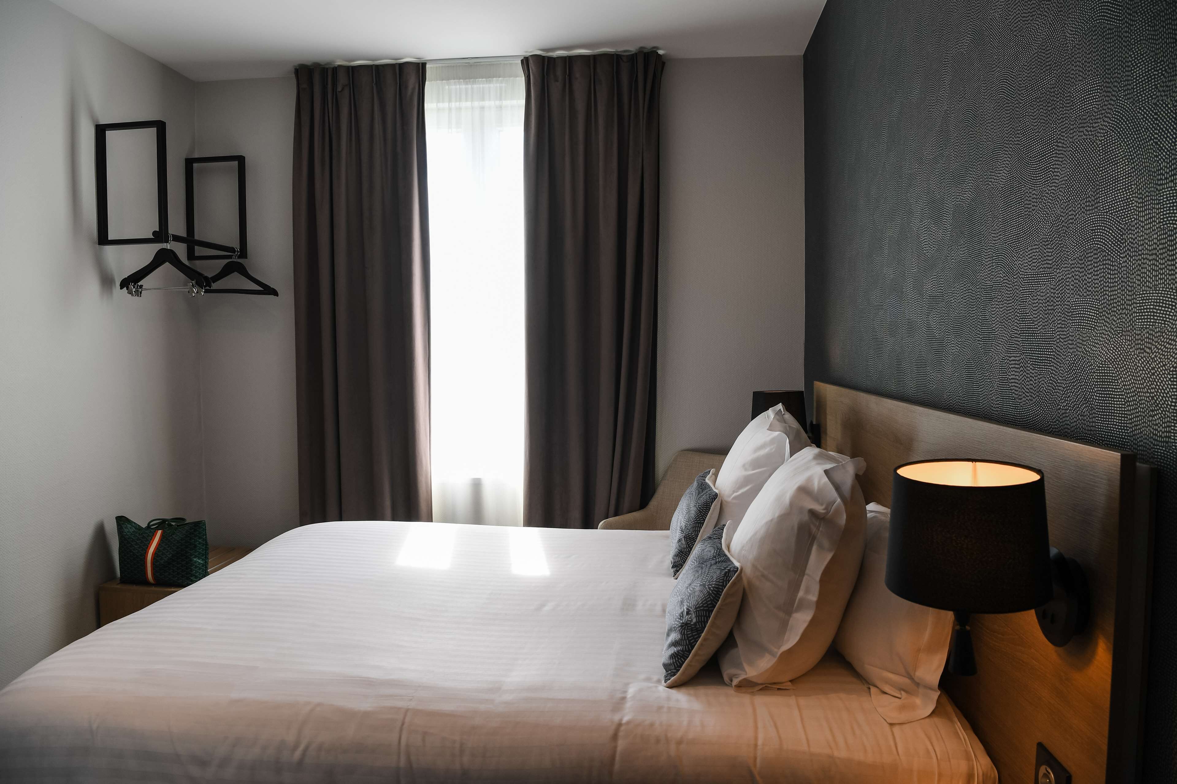 Una camera d'albergo con un letto, un comodino, una lampada, una sedia e una finestra con tende.