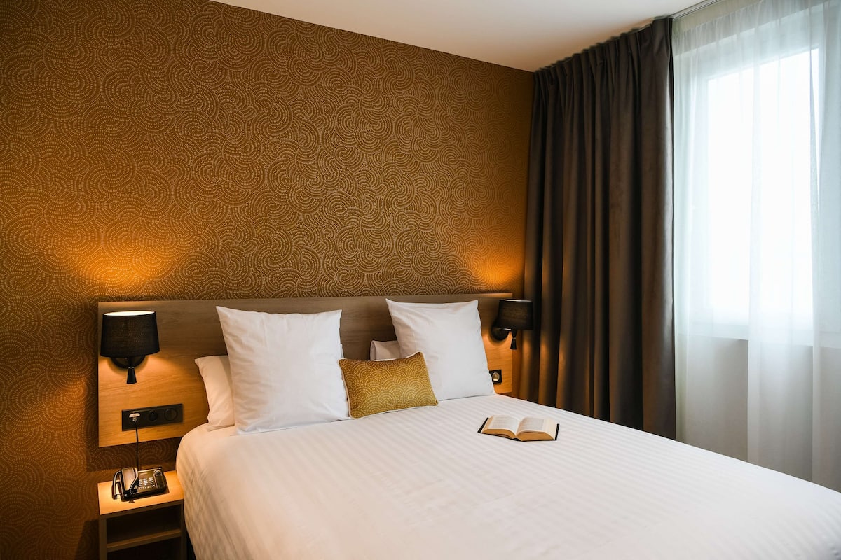 Habitación de hotel con pared estampada, cama con ropa de cama blanca, un libro sobre la cama y mesita de noche con lámpara y teléfono.