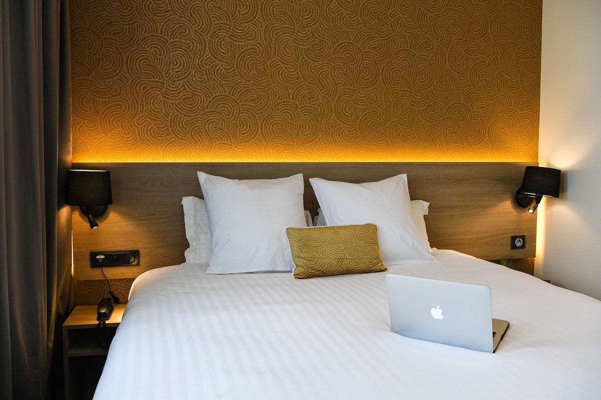 Una cama bien tendida con sábanas blancas, una laptop y una almohada de color mostaza.