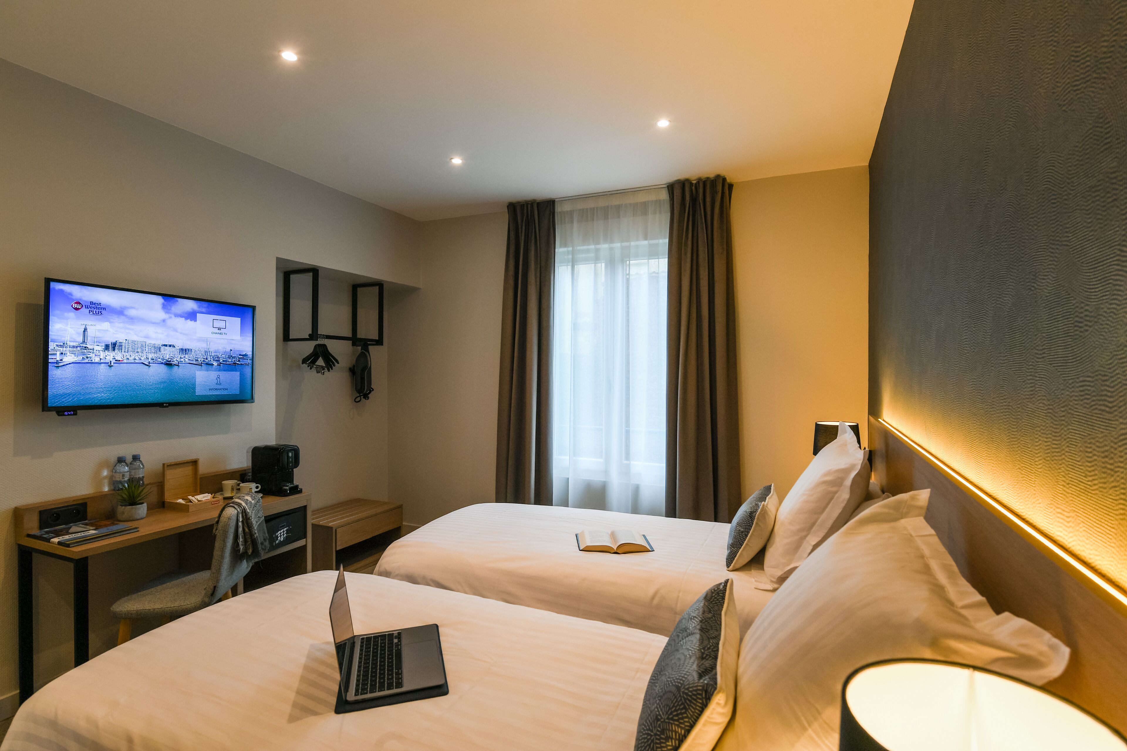 A hotel room with two beds, a desk with a laptop, a television displaying a cityscape, and a large window with curtains.