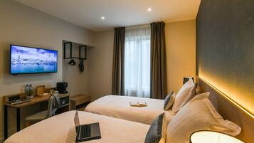 A hotel room with two beds, a desk with a laptop, a television displaying a cityscape, and a large window with curtains.