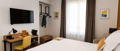 A hotel room with a bed, a desk with a chair, a TV, and a window with curtains.