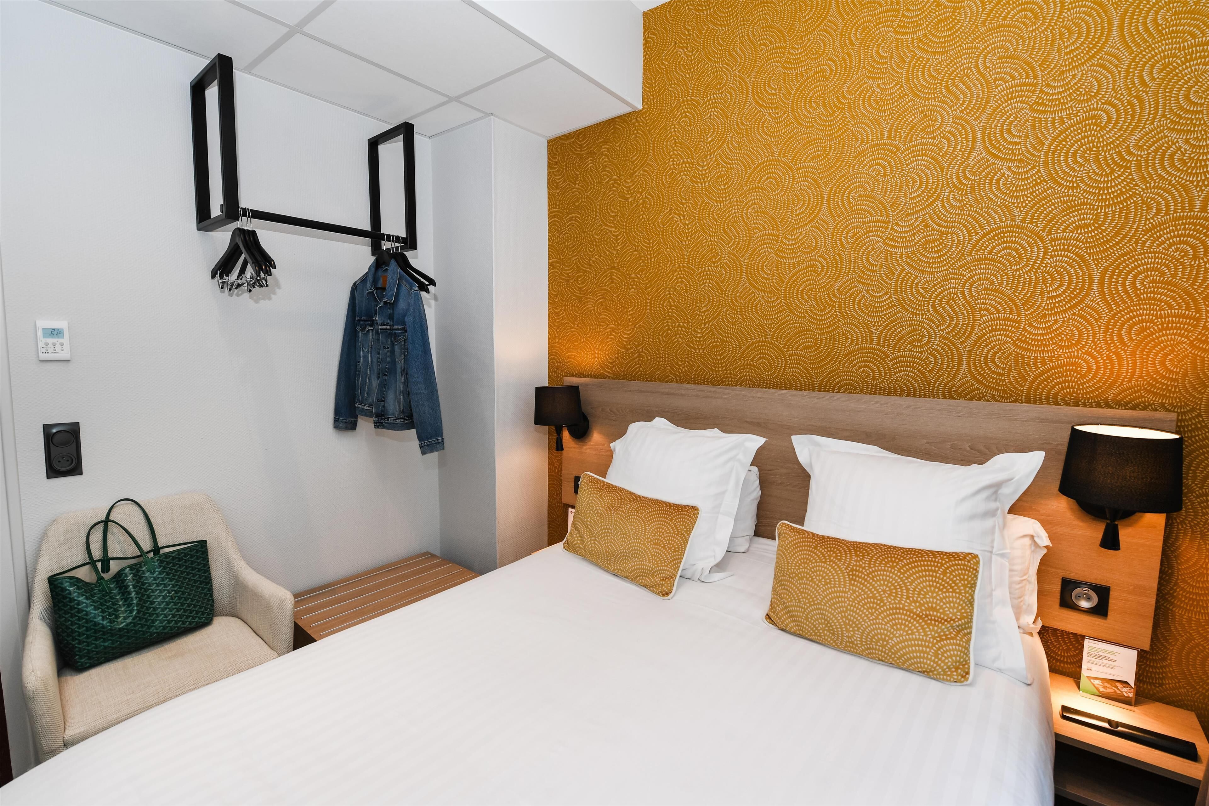 A hotel room with a bed, a chair, a hanging coat rack, and a wall with a patterned wallpaper.