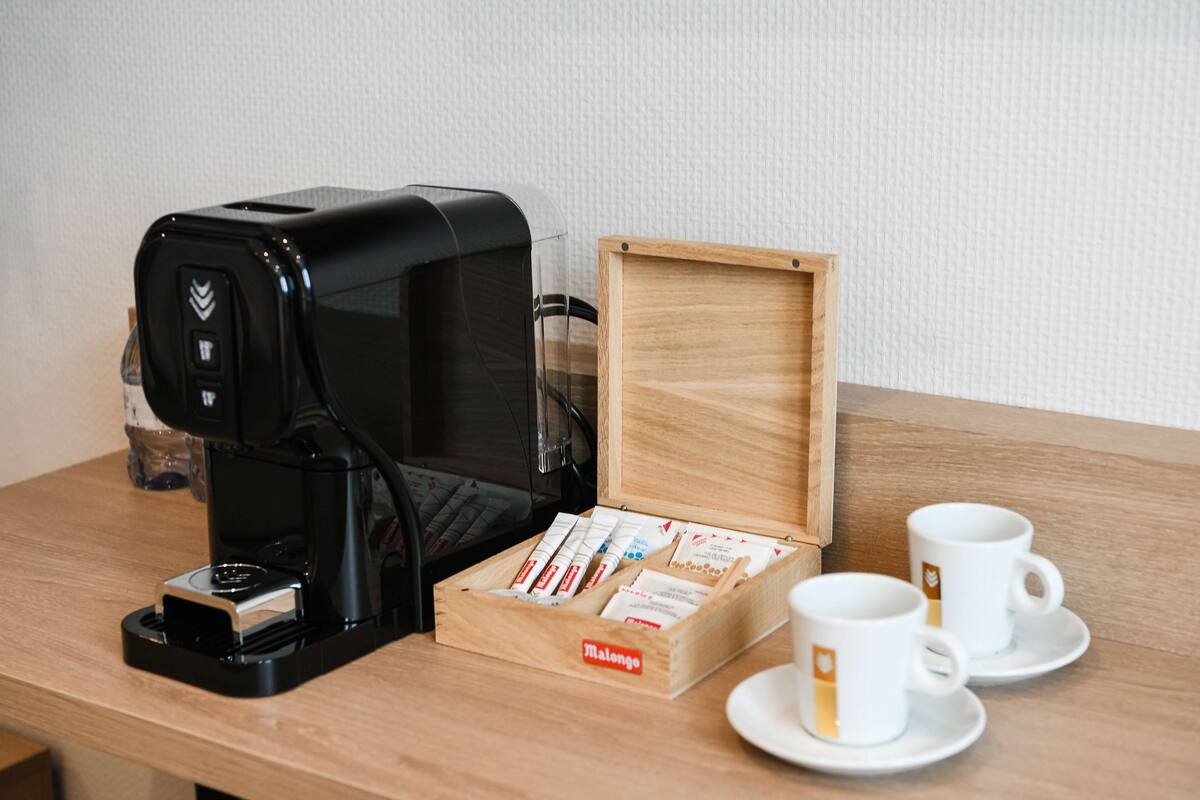 Una cafetera, una caja de cápsulas de café y dos tazas blancas sobre una mesa de madera.