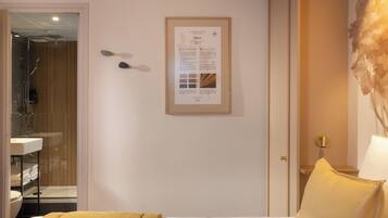 Ein Hotelzimmer mit einem Bett, einem Nachttisch mit Lampe, einem gerahmten Informationsplakat an der Wand und einem durchgegangenes Badezimmer.