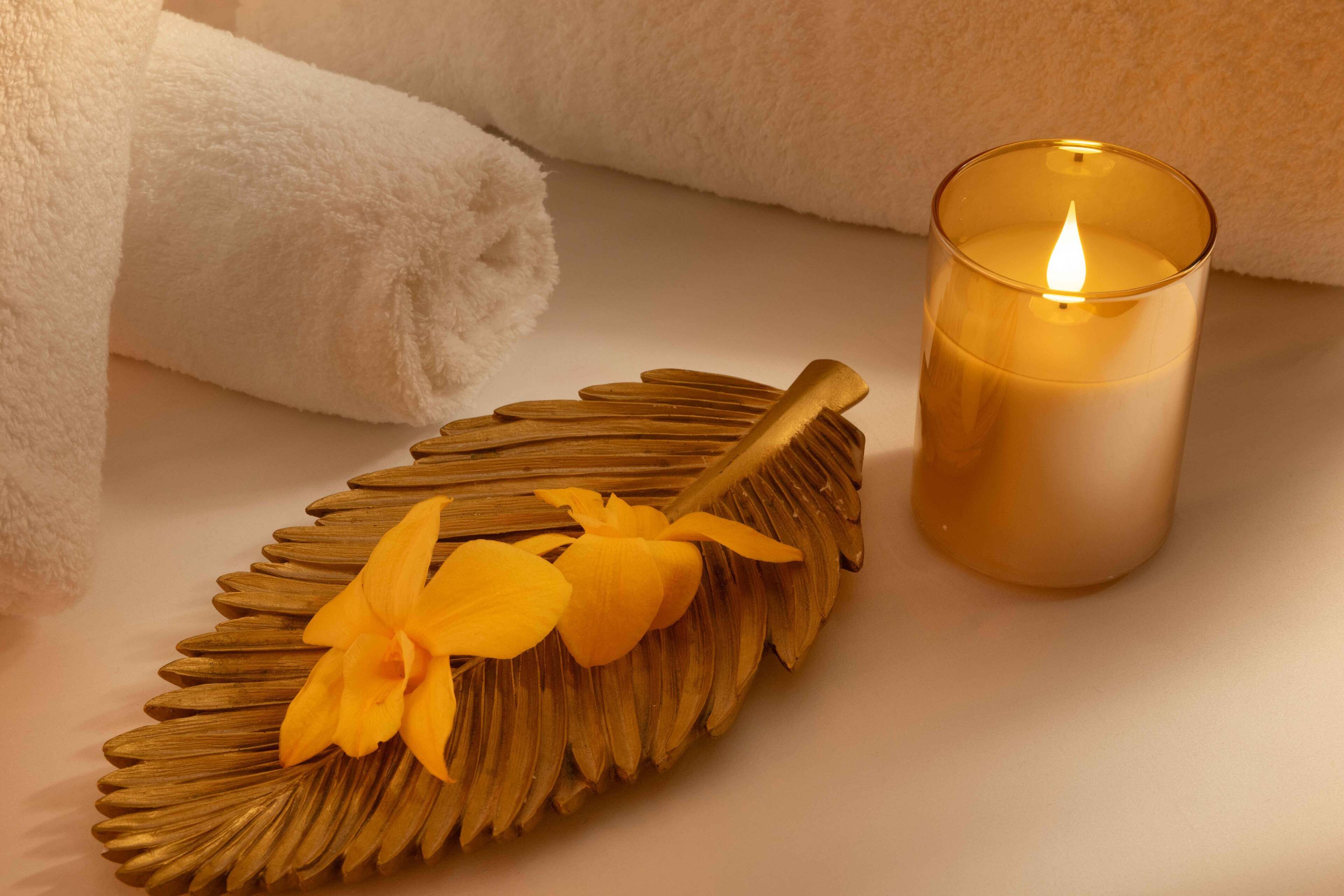 A candle in a glass holder, a rolled white towel, and a decorative leaf with yellow flowers on a white surface.