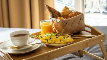 Uma bandeja de café da manhã com uma xícara de café, um copo de suco de laranja, ovos mexidos, croissants e uma cesta de pão sobre uma mesa de madeira.