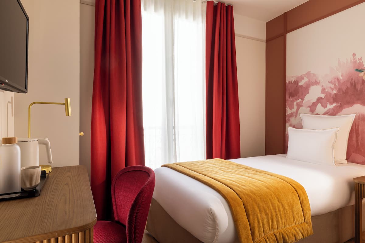 A hotel room with a bed, a desk with a lamp, a chair, and a painting on the wall.