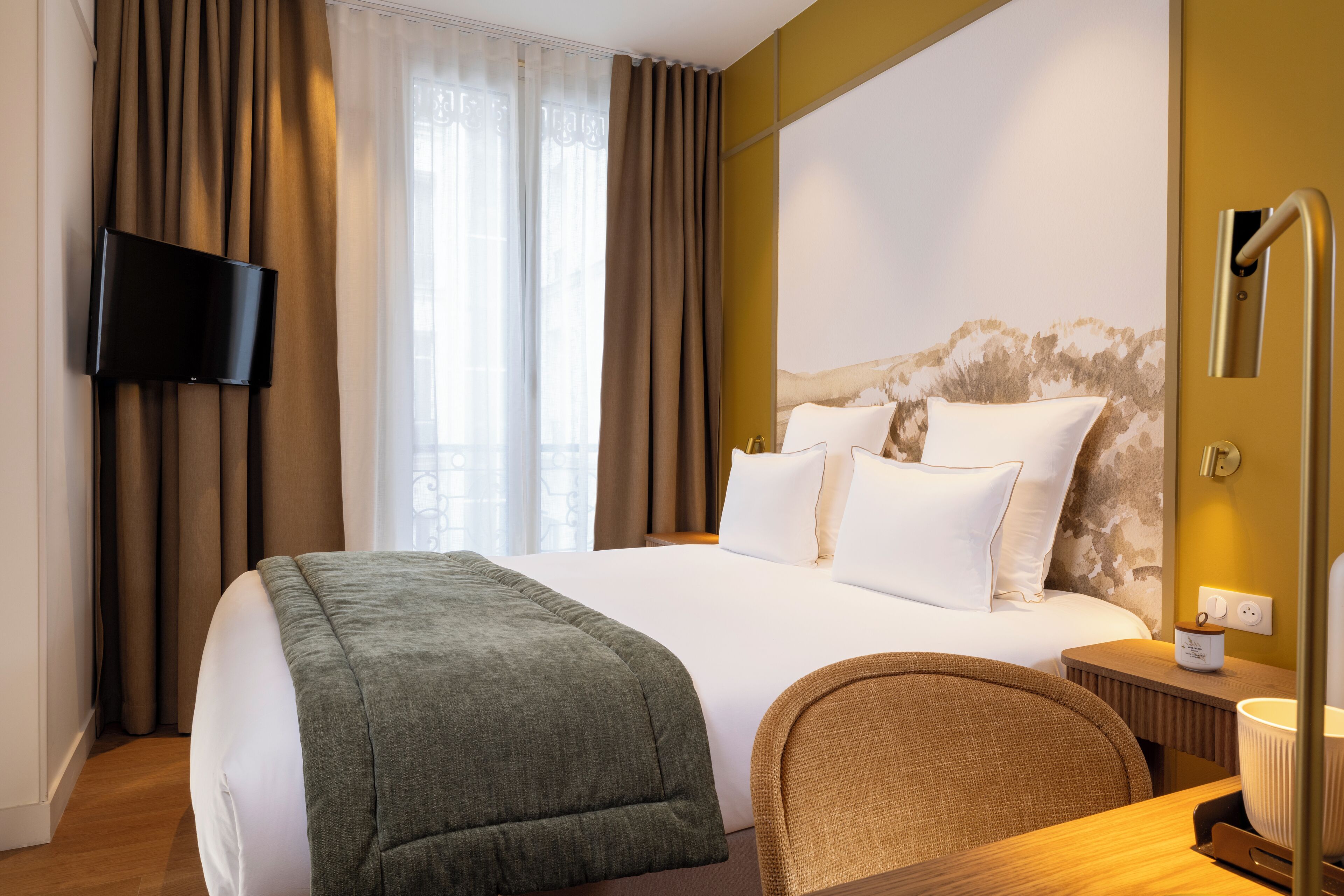 A hotel room with a large bed, a flat-screen TV mounted on the wall, a bedside table with a lamp, and a view of the outdoors through the window.