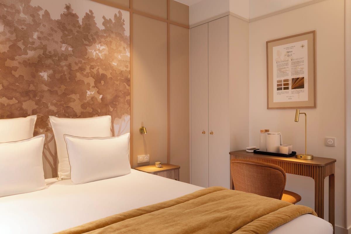 A hotel room with a large bed, a desk with a chair, a framed informational poster, and a small table with a lamp and some items.