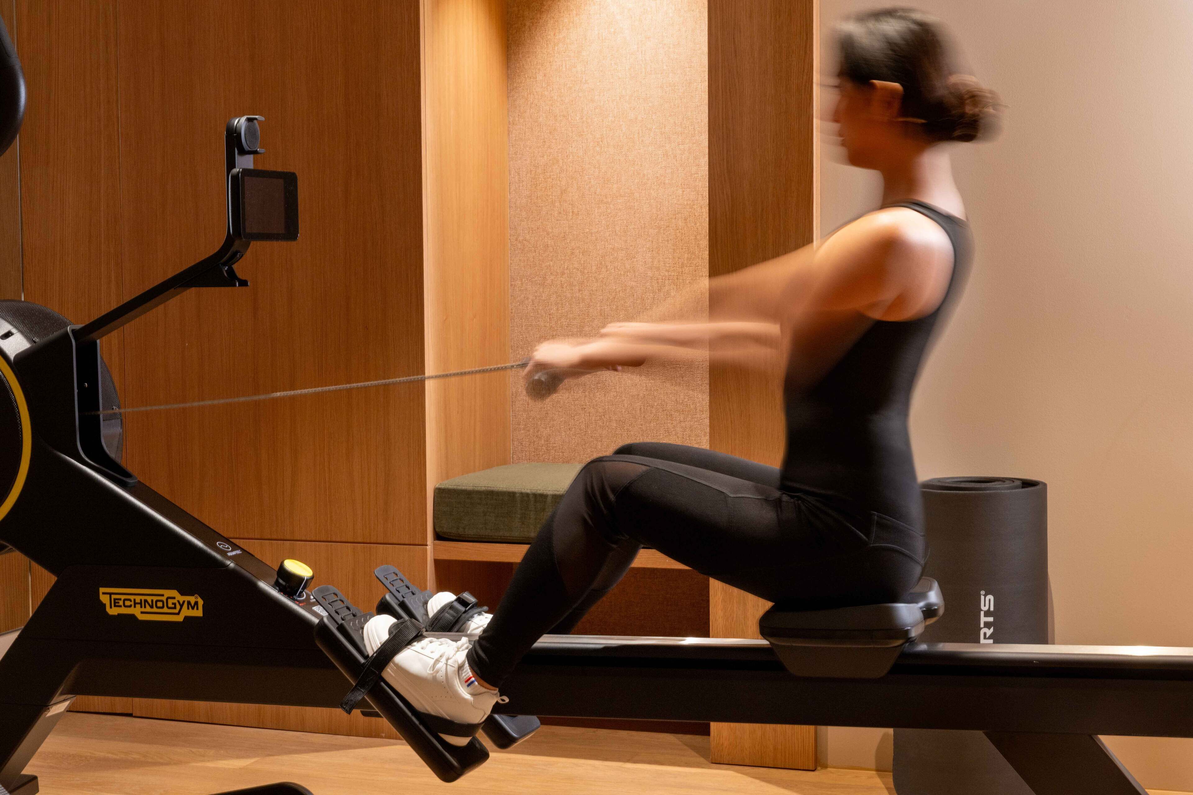A person using a rowing machine in a room with wooden walls and a bench.