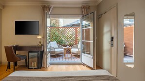 A bedroom with a bed, a desk, a chair, and a small table with a lamp. There is a sliding glass door leading to a balcony with a wooden deck and a lattice fence. - Hôtel Le Walt by Inwood Hotels (Paris)
