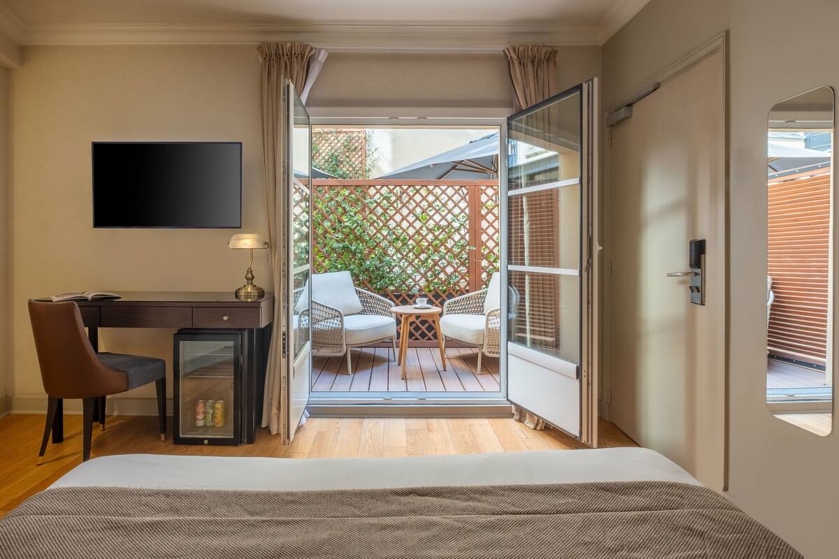A bedroom with a bed, a desk, a chair, and a small table with a lamp. There is a sliding glass door leading to a balcony with a wooden deck and a lattice fence.