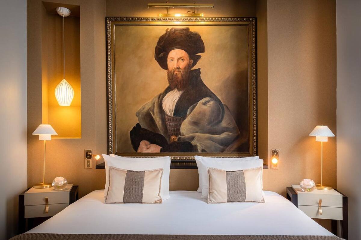 A hotel room with a large portrait on the wall, two bedside lamps, and a bed with white bedding and two pillows.