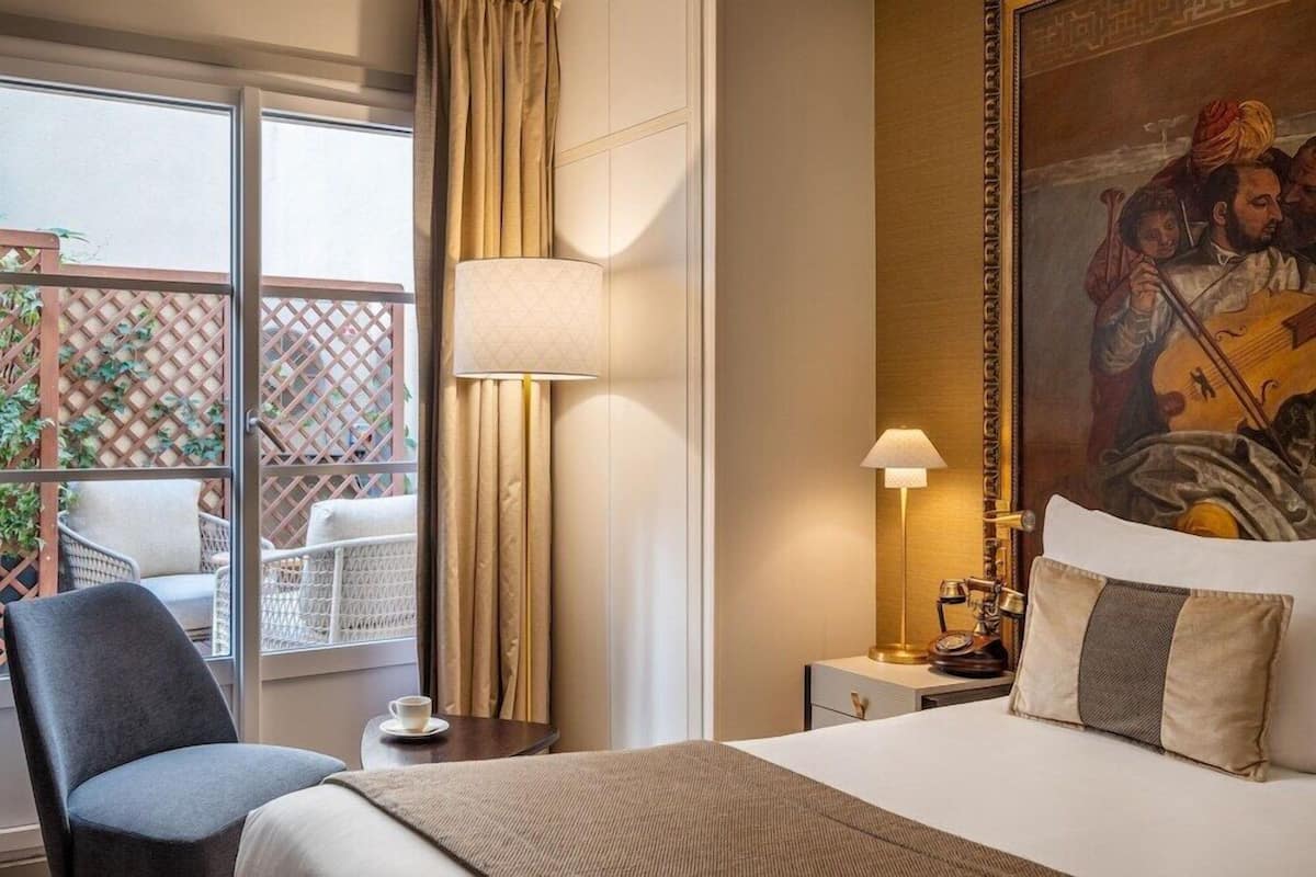 A hotel room with a bed, a chair, a nightstand, a lamp, a painting, and a view through a window with a lattice.
