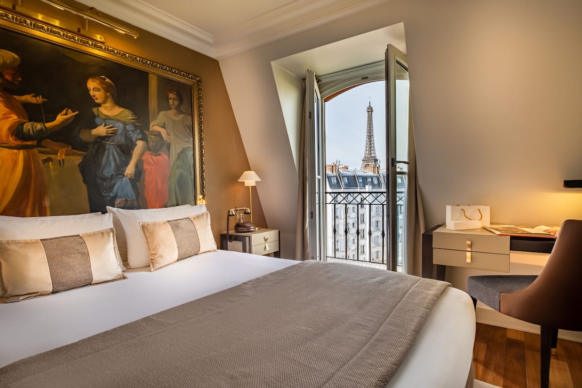 A hotel room with a large bed, a desk, a chair, a lamp, and a painting of a historical scene.