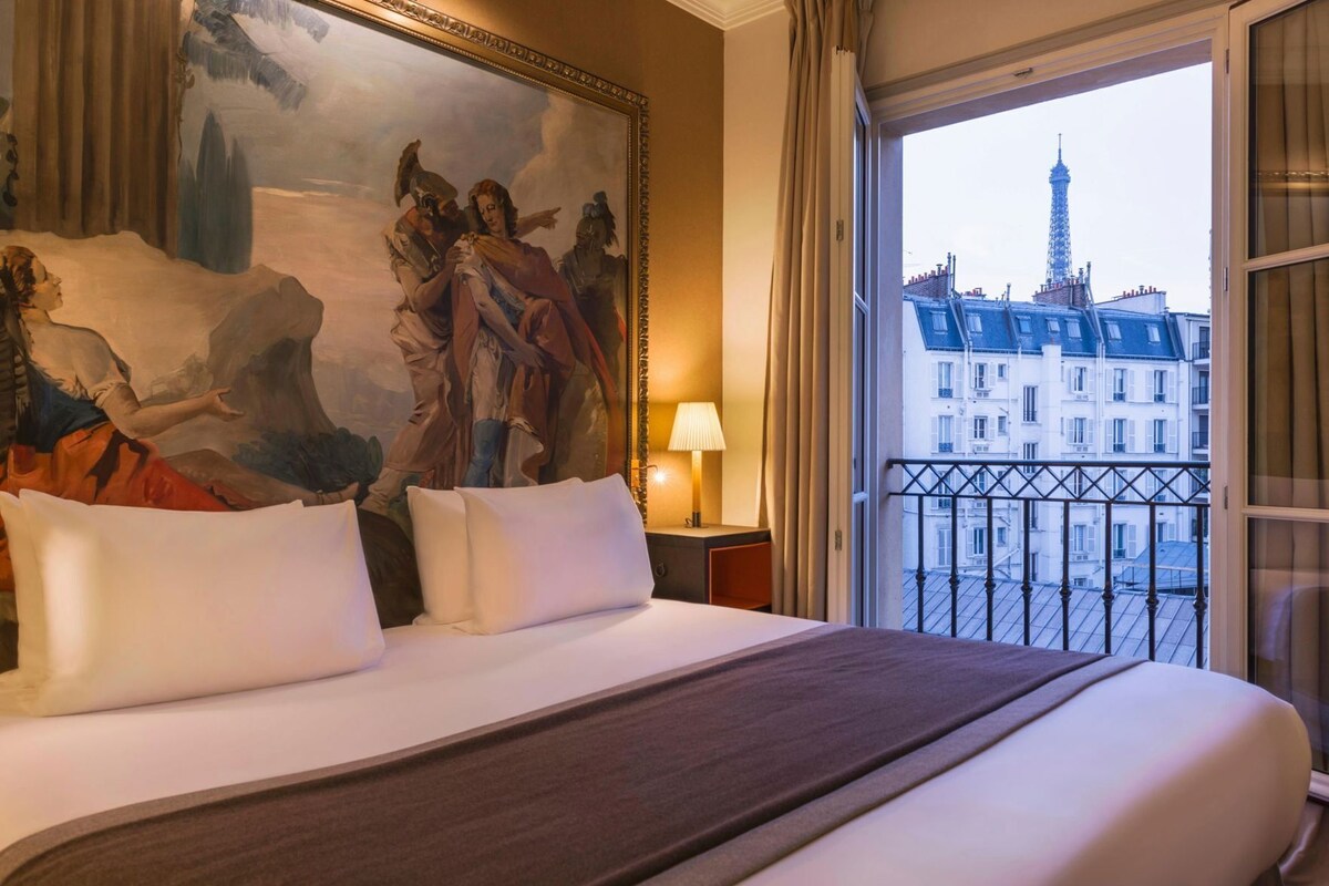A hotel room with a large bed, a painting on the wall, a view of the Eiffel Tower, and a balcony.
