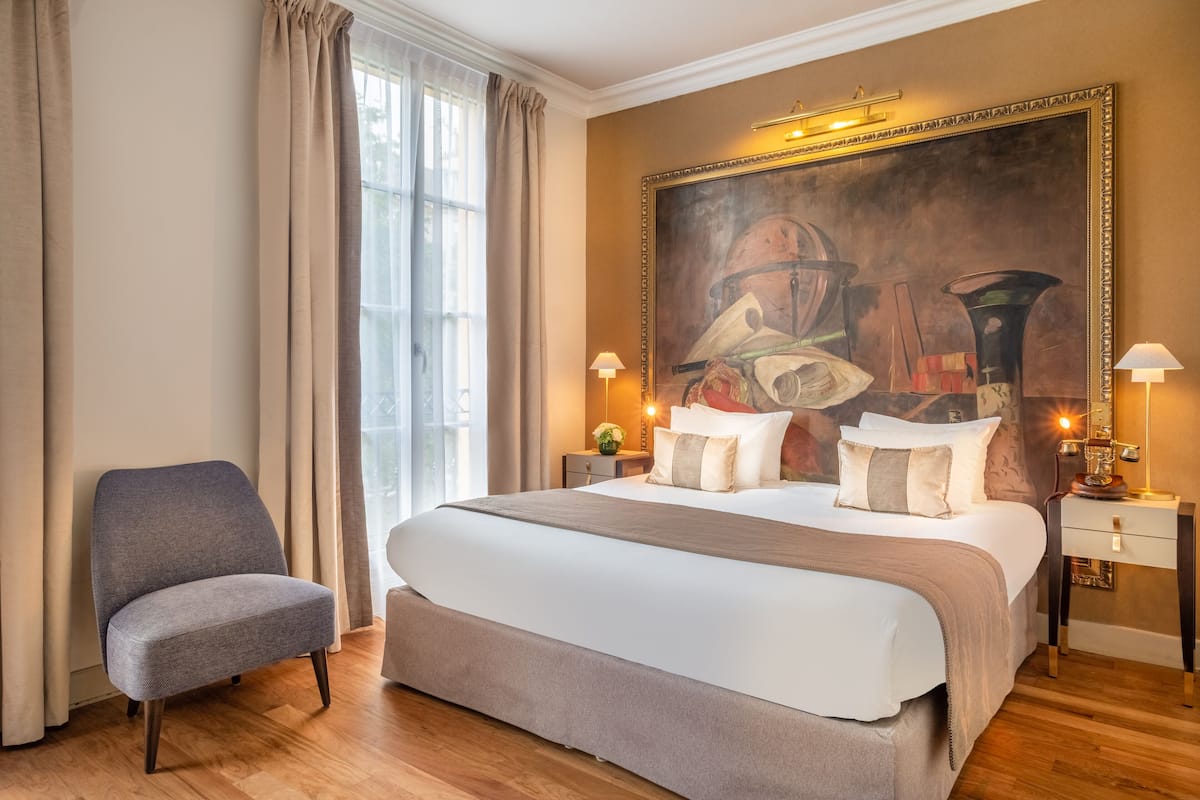 A hotel room with a large bed, a chair, a nightstand with a lamp, and a painting on the wall.