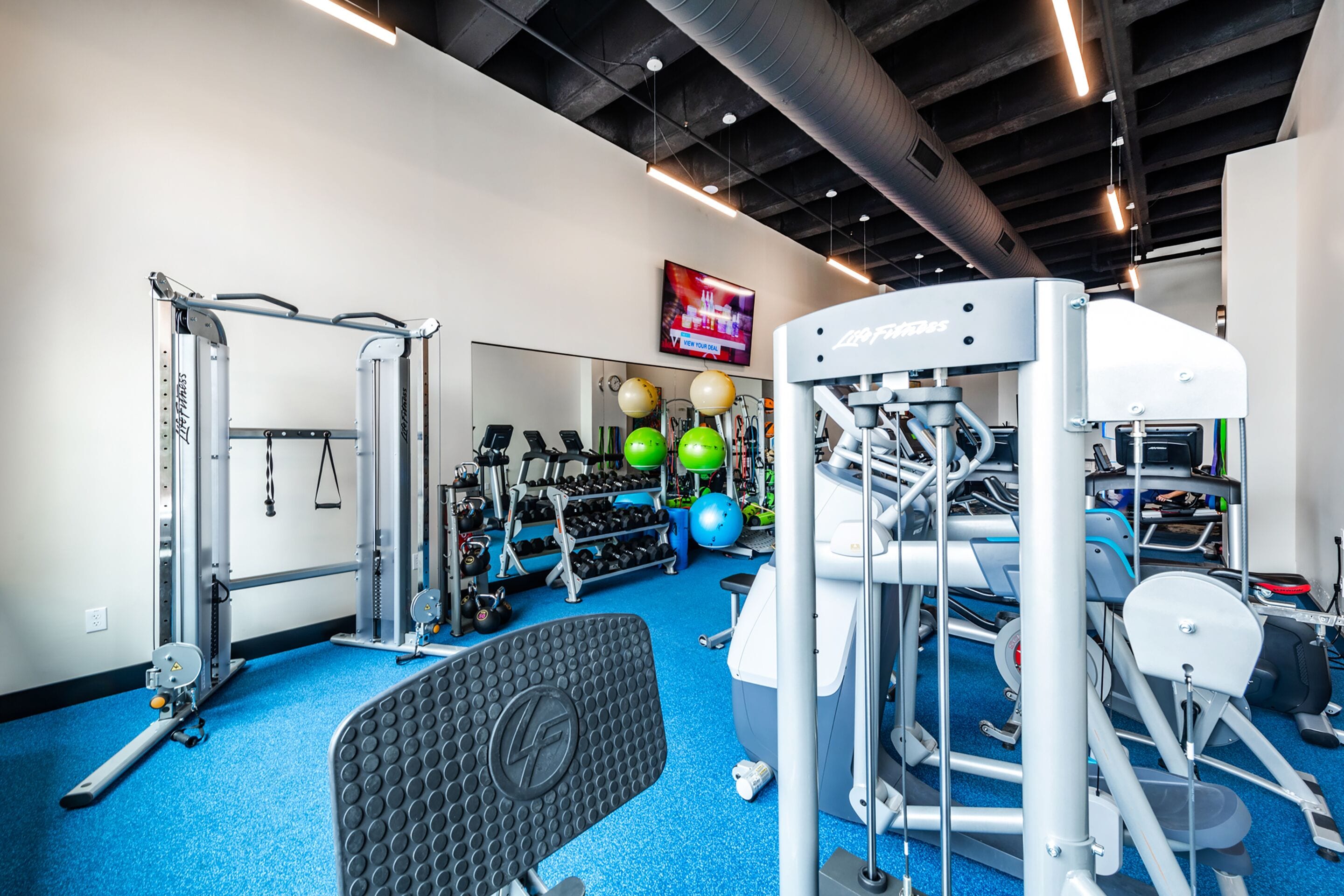 A gym with various exercise equipment, a mirrored wall, and a television displaying a news channel.