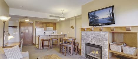 Una sala de estar con chimenea, un televisor montado arriba y una cocina al fondo.