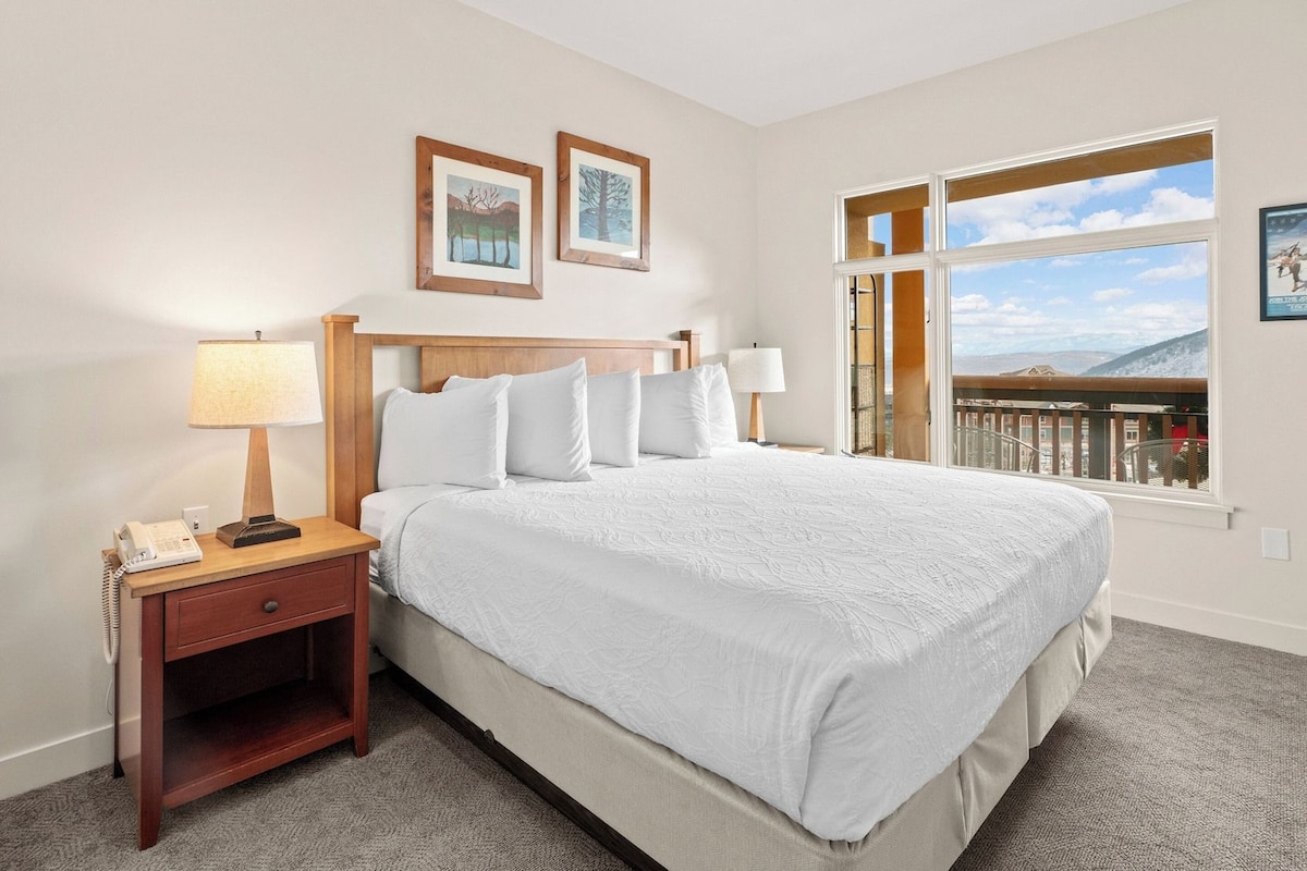 Habitación de hotel con una cama grande, dos lámparas de noche, una mesita con teléfono, ventana con vista a las montañas y dos cuadros en la pared.