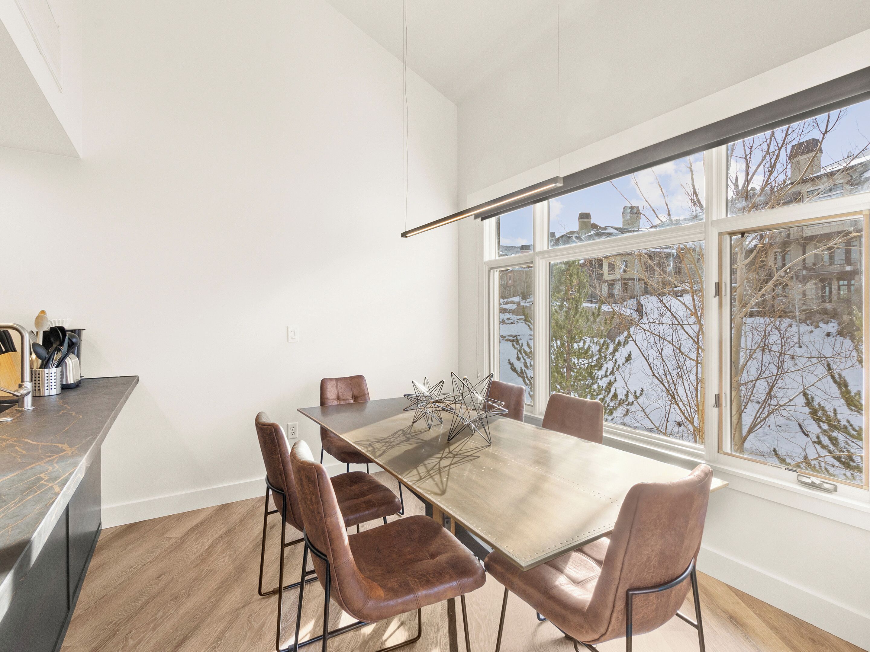Un espace repas moderne avec une grande table et six chaises, un comptoir de cuisine équipé d’ustensiles et une vue sur un paysage enneigé grâce à de grandes fenêtres.