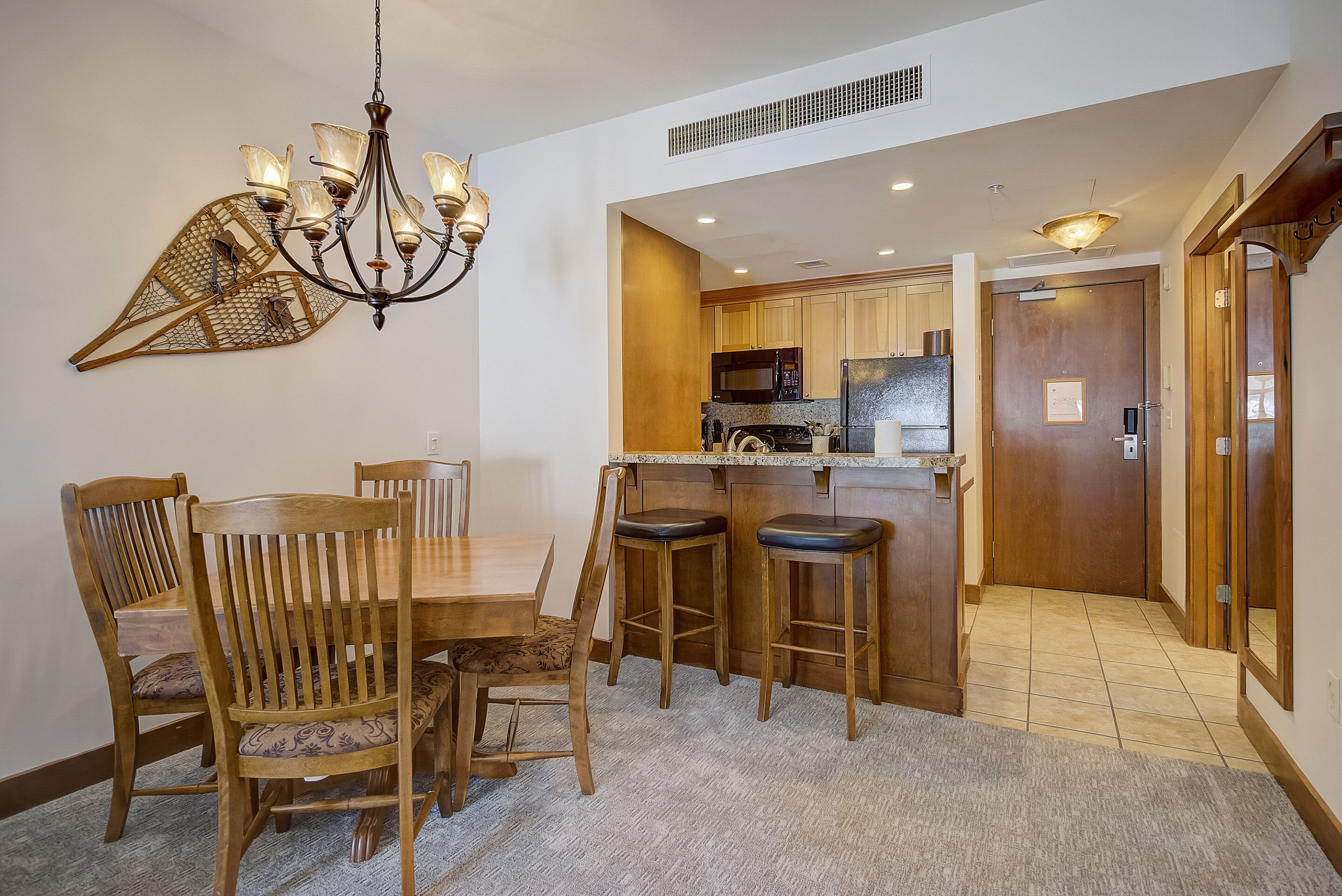 A dining area with a wooden table and chairs, a chandelier, and a kitchen in the background.