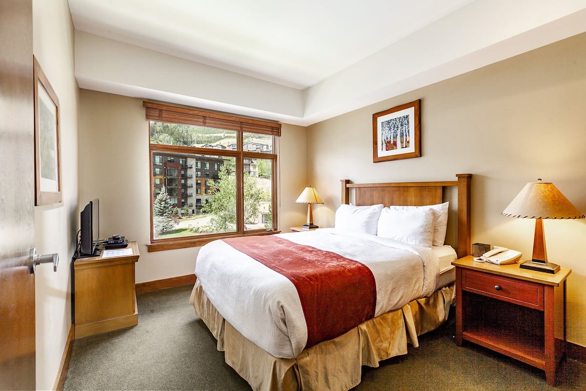Een hotelkamer met een groot bed, twee bedlampen, een houten nachtkastje, een televisie en een raam met uitzicht op gebouwen en bomen.