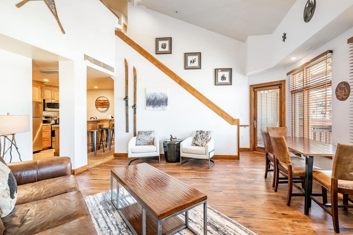 Een ruime woonkamer met een houten salontafel, een leren bank en een eettafel met stoelen. Een trap met houten leuningen leidt naar een hoger niveau.