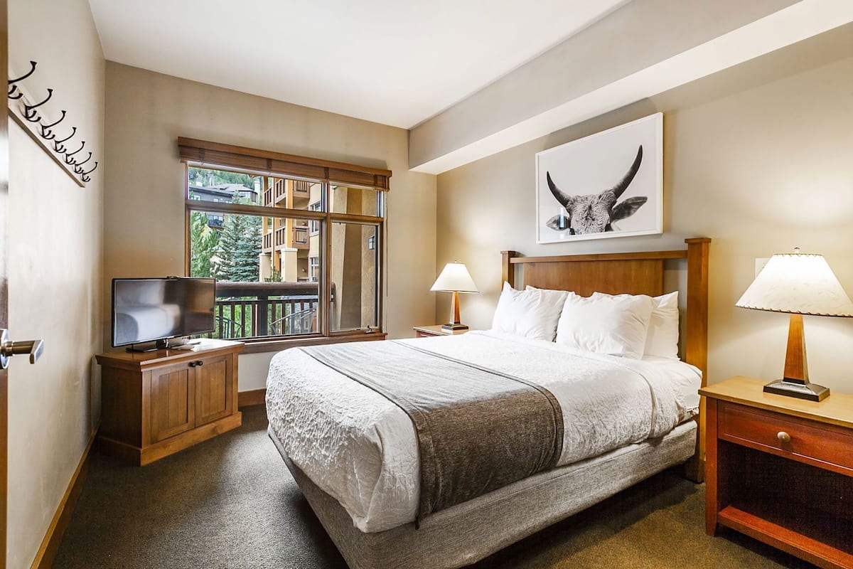 Hotelkamer met een groot bed, twee nachtkastjes met lampen, een televisie op een houten kast, een raam met uitzicht op gebouwen, en een kunstwerk van een stierenkop aan de muur.