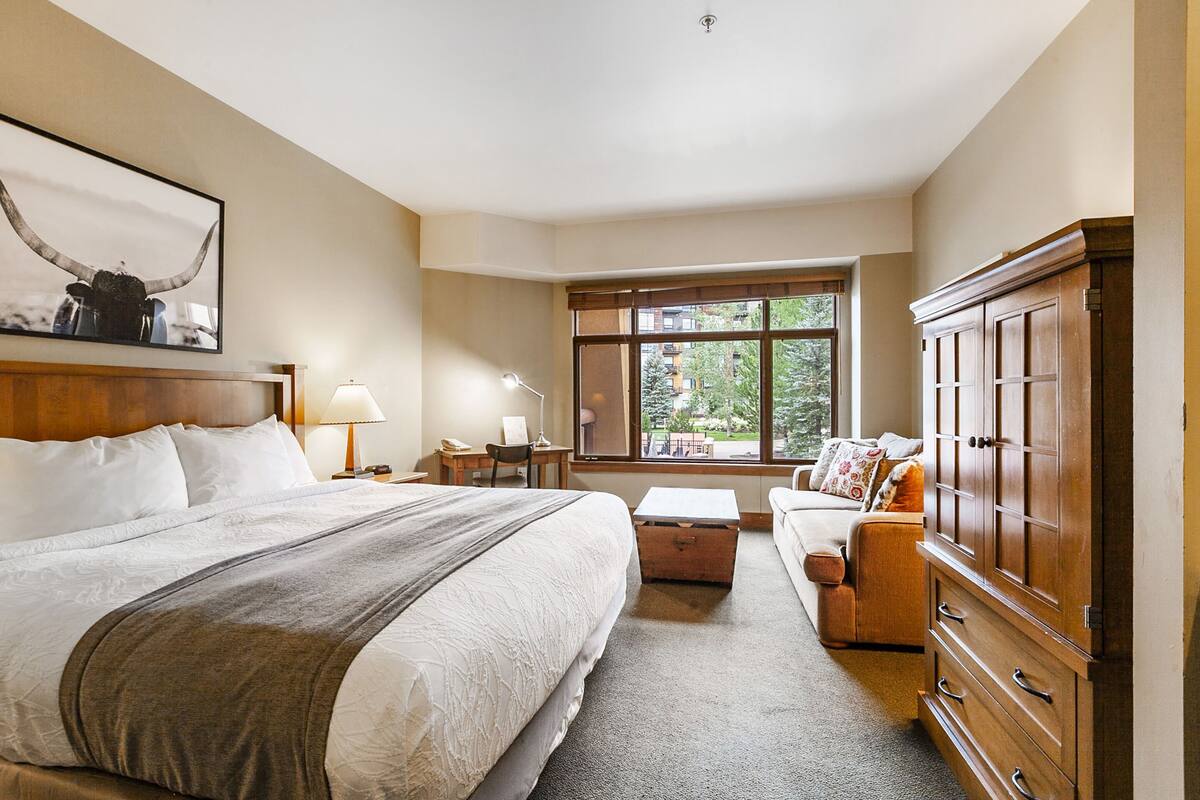Een hotelkamer met een groot bed, een houten garderobe, een bureau met lamp, een raam met uitzicht op groen, en een bank.