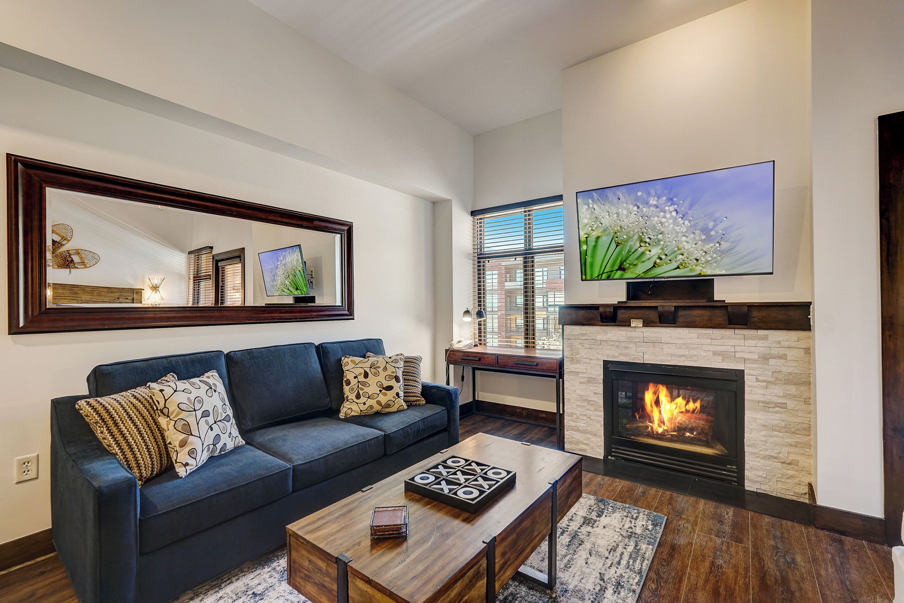 A living room with a fireplace, a large flat-screen TV mounted above it, a dark blue sofa with patterned cushions, and a wooden coffee table.