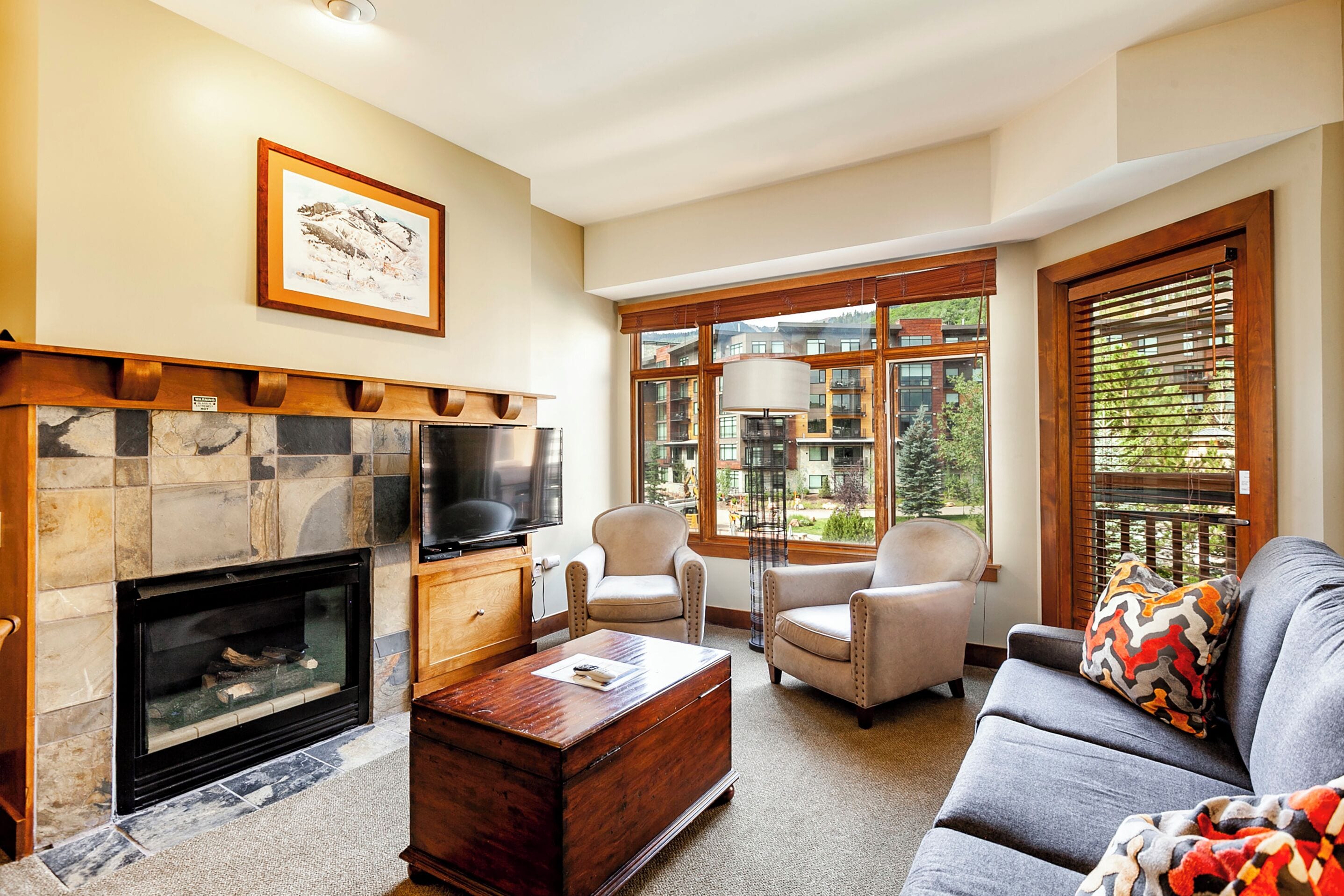 A living room with a stone fireplace, a television, two armchairs, a sofa, and large windows offering a view of buildings and trees.