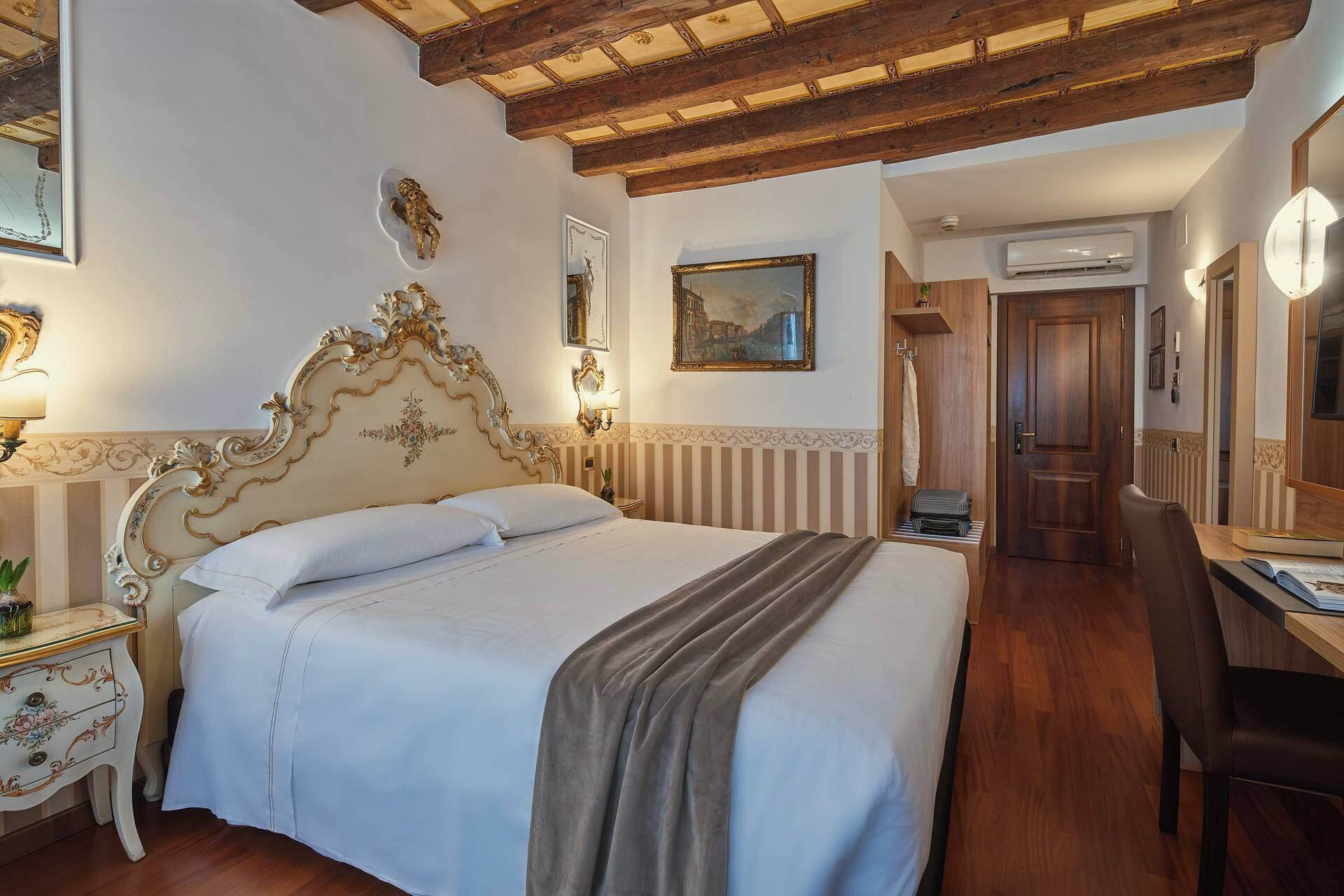 Een hotelkamer met een groot bed, een houten plafond, een bureau met stoel, en een deur naar een andere kamer.