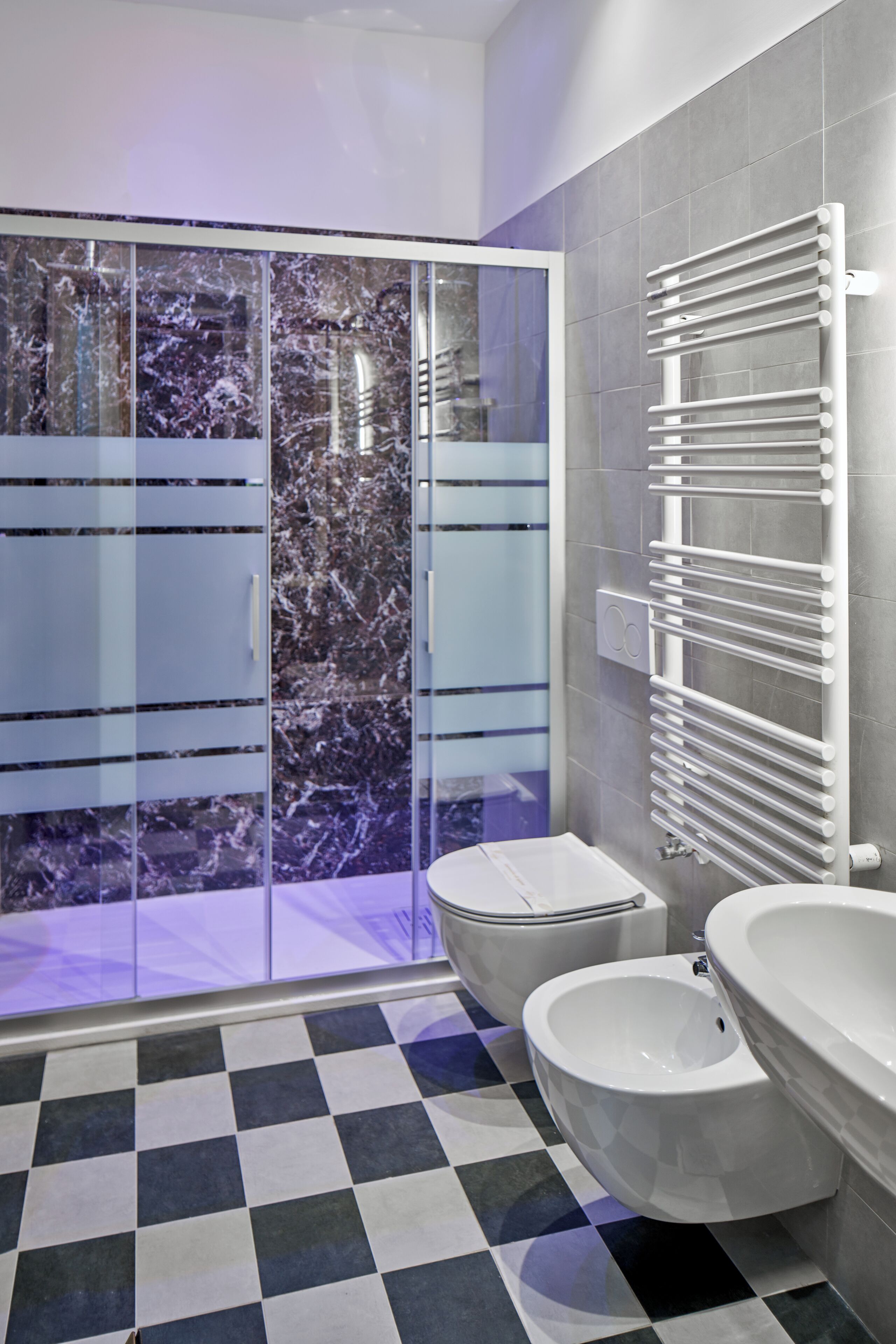 Un bagno moderno con pavimento a scacchi, box doccia in vetro, un water bianco e un bidet bianco.