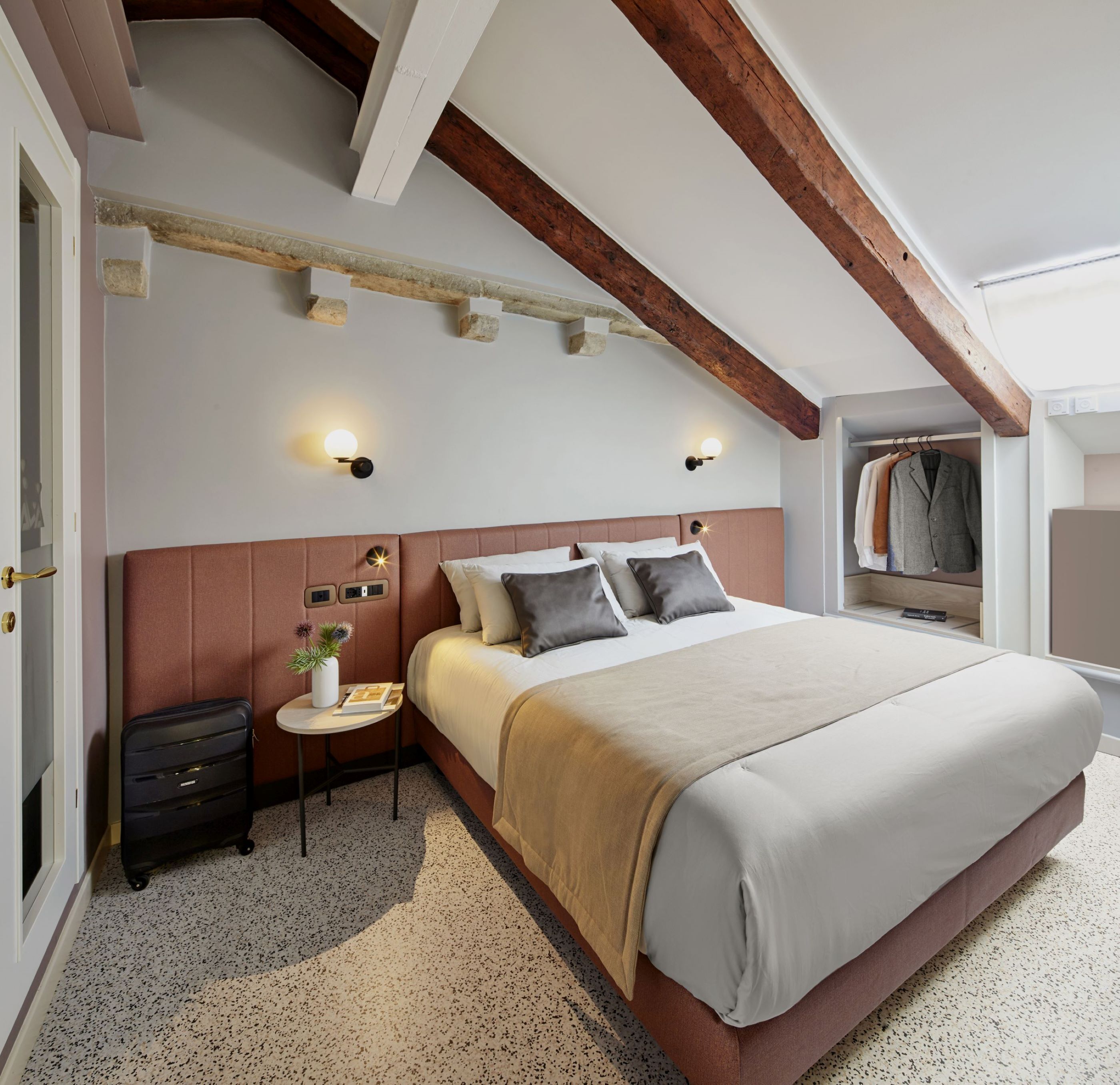 A bedroom with a large bed, a nightstand, a small table with a plant, and a closet with hanging clothes.