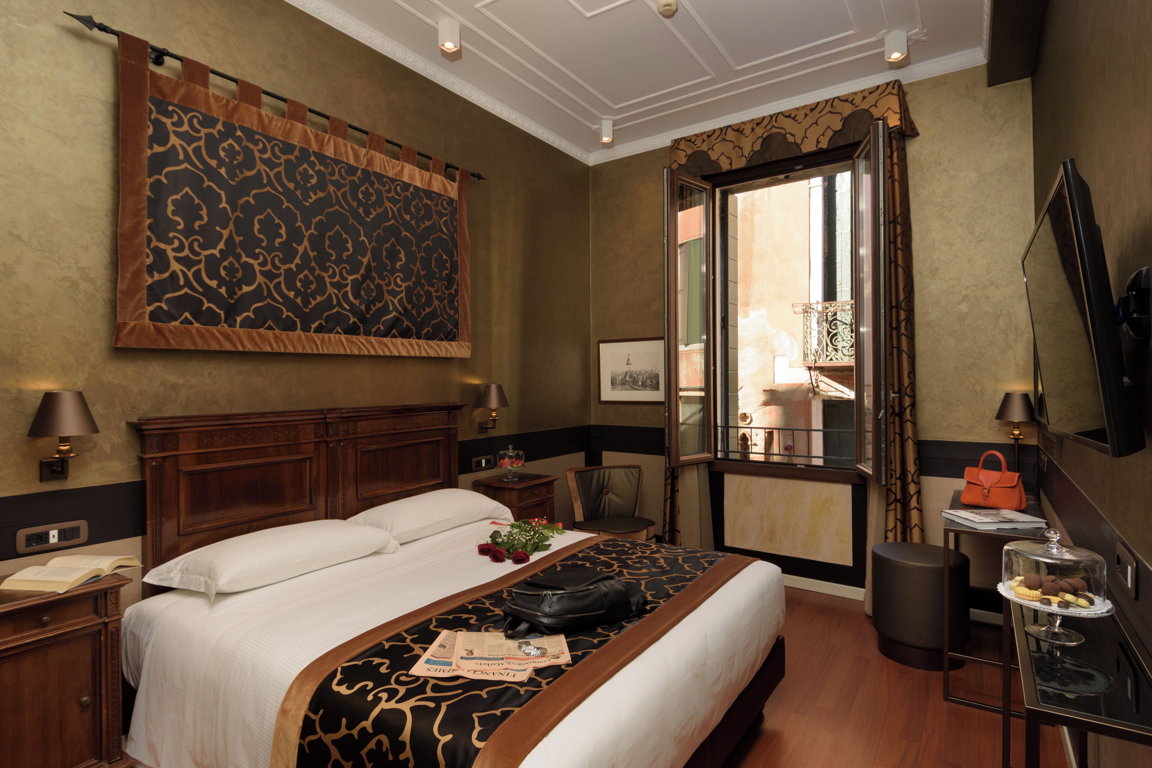A hotel room with a large bed, a desk with a lamp, a chair, a small table with a glass display, and a large window with curtains.