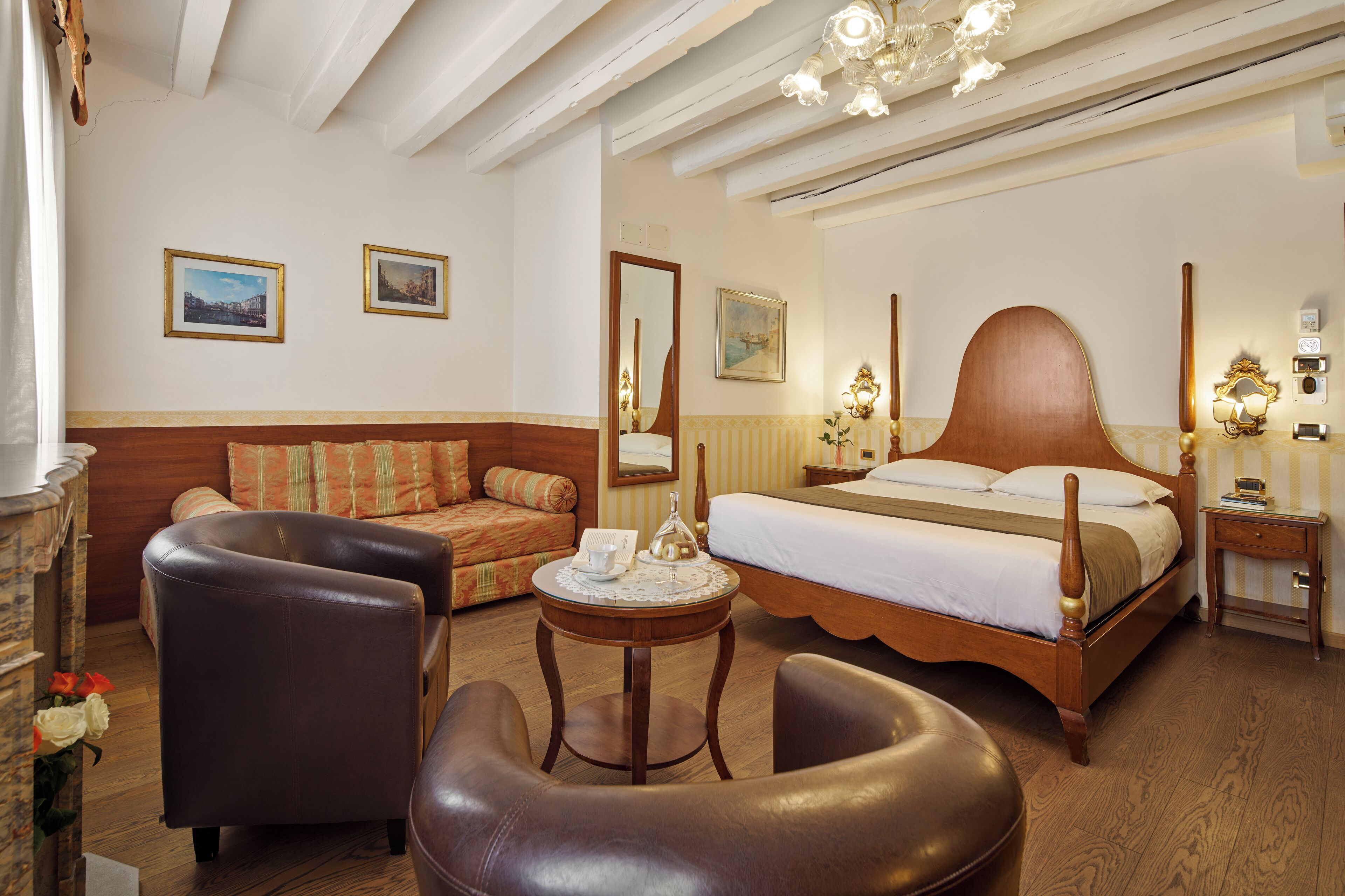 A hotel room with a wooden bed, a sofa, two armchairs, a small round table with a lamp, and a mirror on the wall.