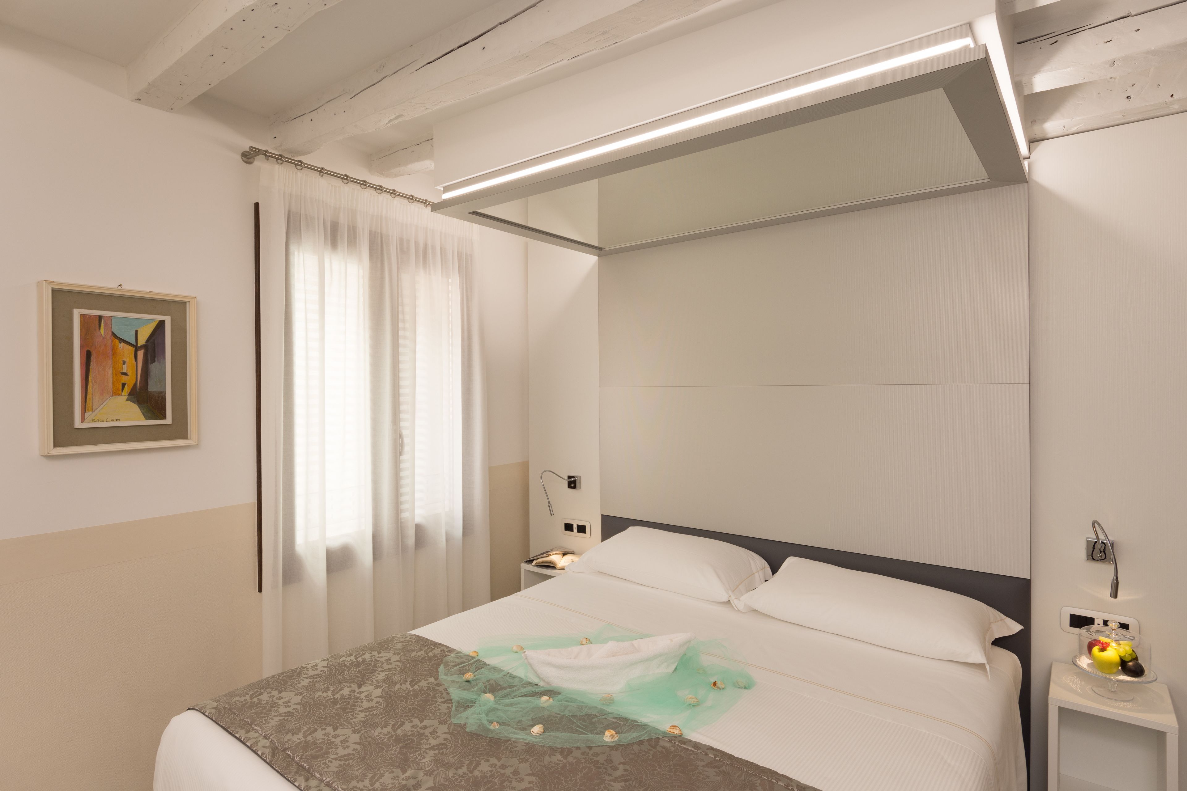 A hotel room with a bed, a bedside table with a lamp, a painting on the wall, and a window with curtains.