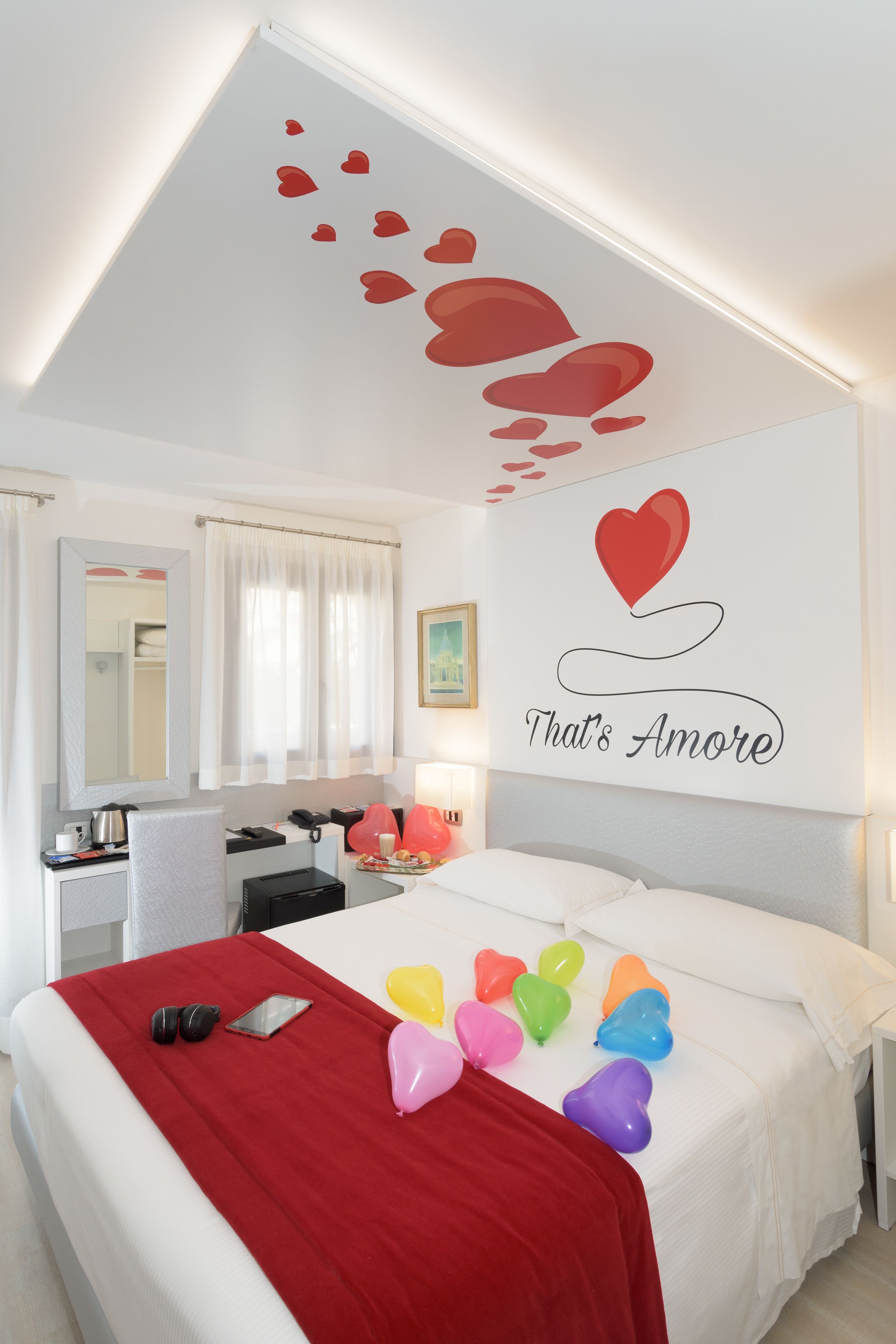 A hotel room with a bed, a desk, a chair, a mirror, and a wall with a heart decoration and the phrase "That's Amore".