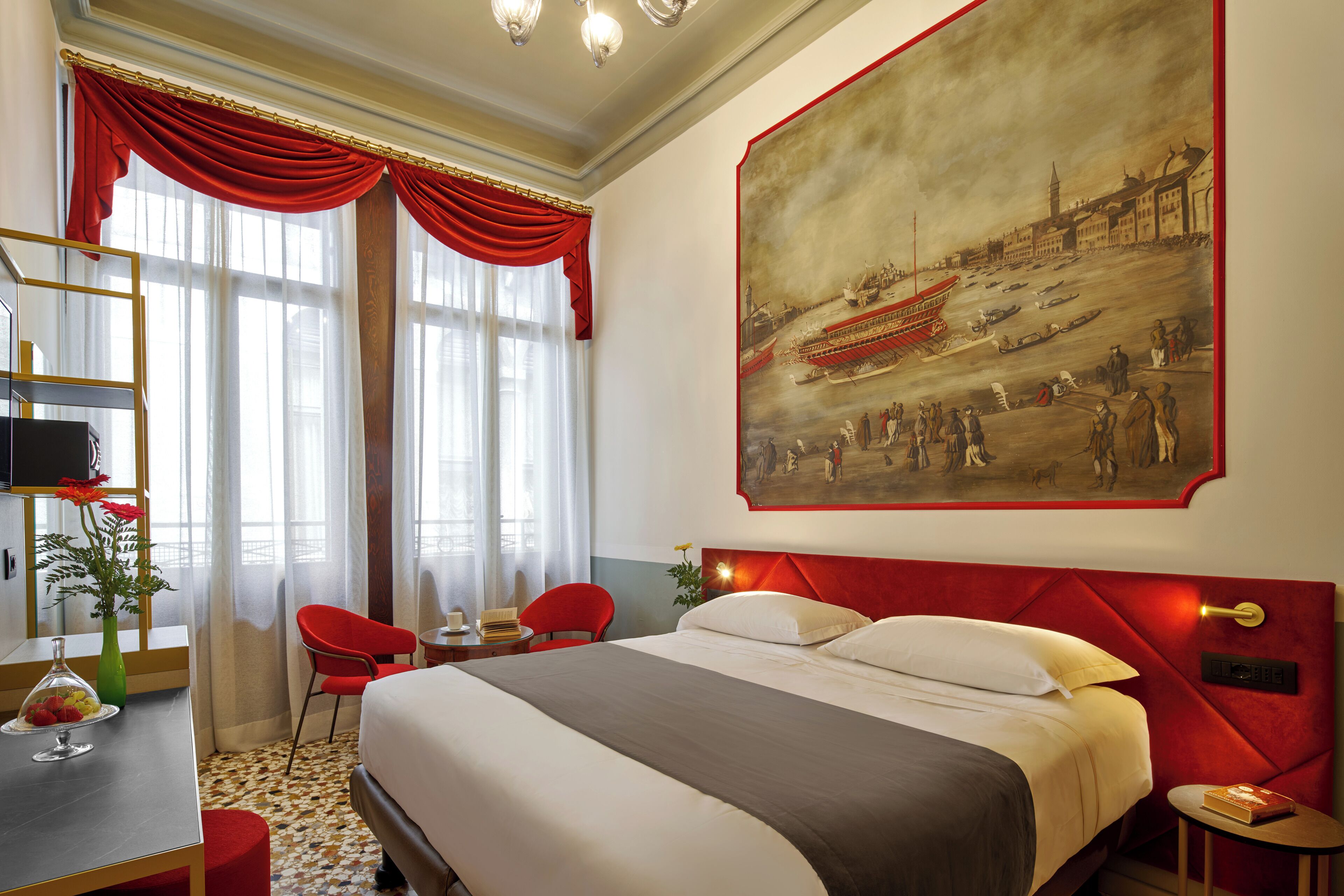 A hotel room with a large bed, red curtains, a painting of a historical scene, a desk with a vase of flowers, and a red chair.
