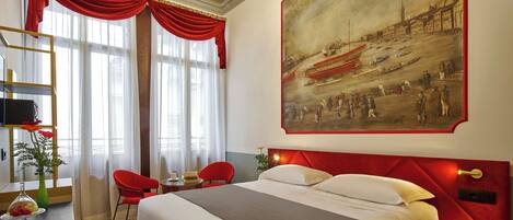 Une chambre d’hôtel avec un grand lit, des rideaux rouges, un tableau représentant une scène historique, un bureau agrémenté d’un vase de fleurs et un fauteuil rouge.