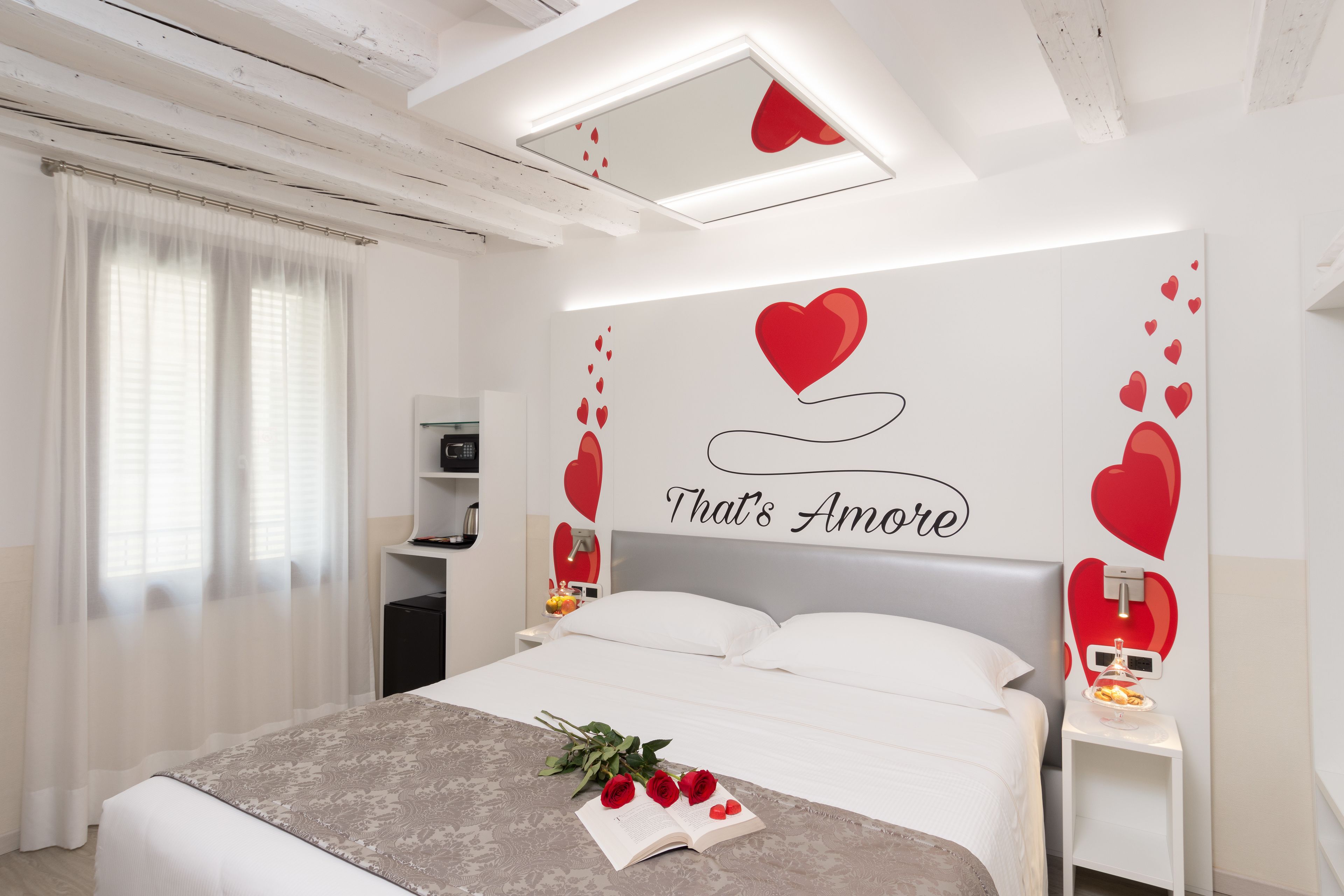 A modern bedroom with a bed, a bedside table, a nightstand, a wall with heart decorations, and a window with curtains.