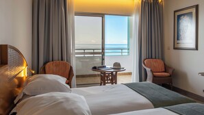 A hotel room with a bed, two chairs, a small table with a hat, and a view of the sea through a sliding door. - Pestana Ocean Bay Resort (Funchal)