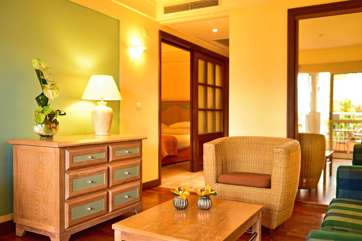 Une chambre d’hôtel avec une commode en bois, une table ornée de deux vases, un fauteuil en osier et un lit en arrière-plan.